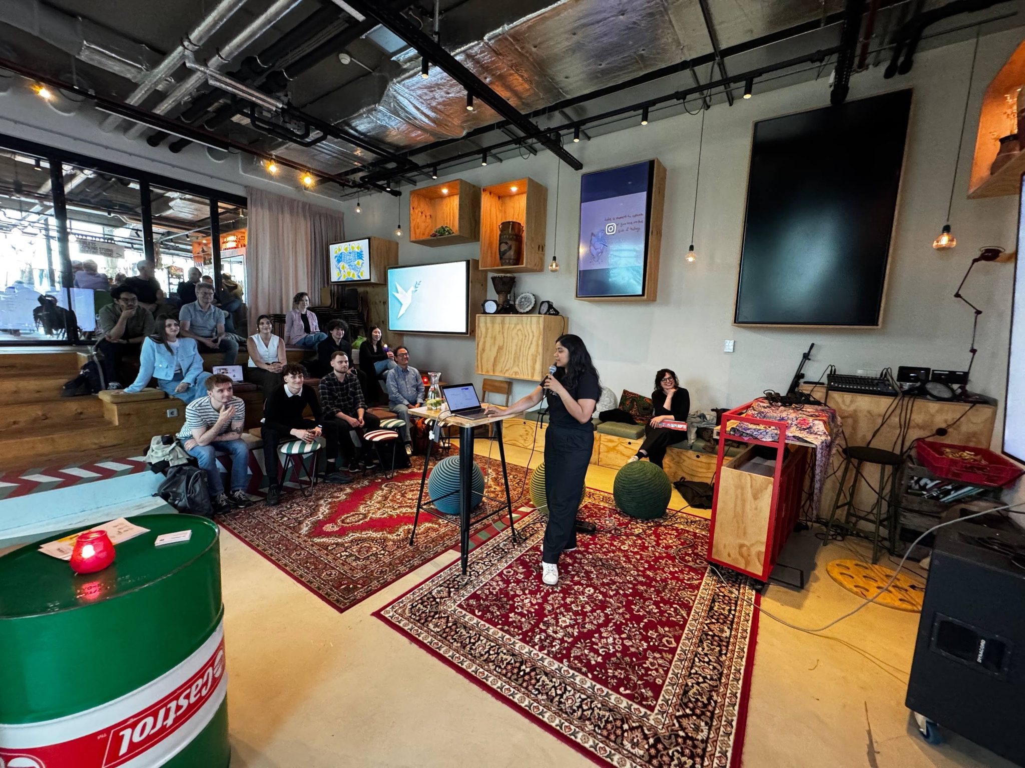 A presenter speaking to attendees seated in an informal venue at the Vibe Coding Open Mic in Amsterdam.